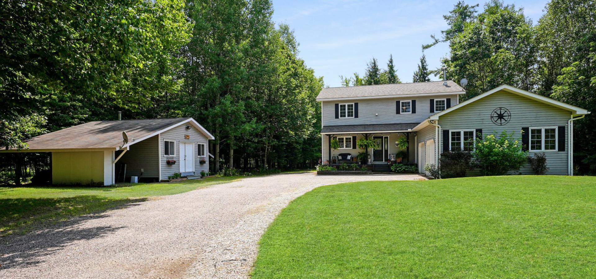 Grey house with attached garage, green grass, gravel laneway, and detached shop