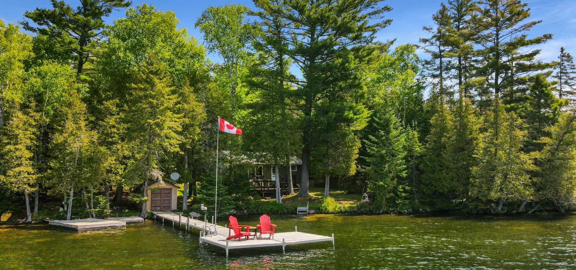 Cottage hidden behind trees, 2 red muskoka chairs on a dock with canada flag.