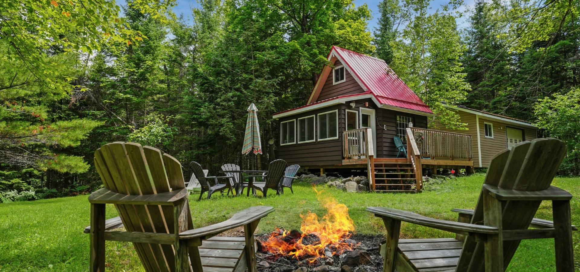 Cabin with red roof and 2 chairs sitting in front of a fire