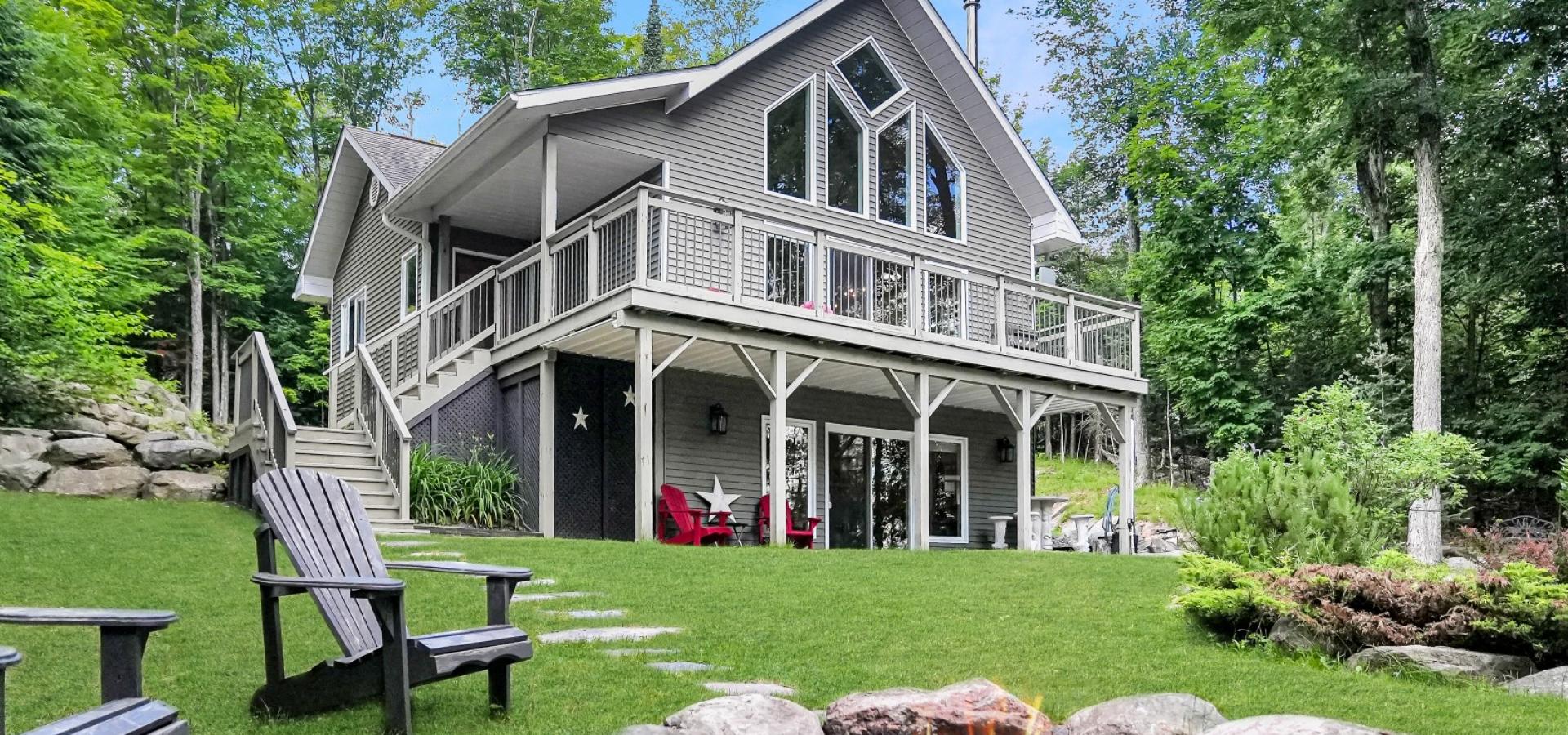grey chalet style house with wrap around upper deck, and firepith with 2 muskoka chairs