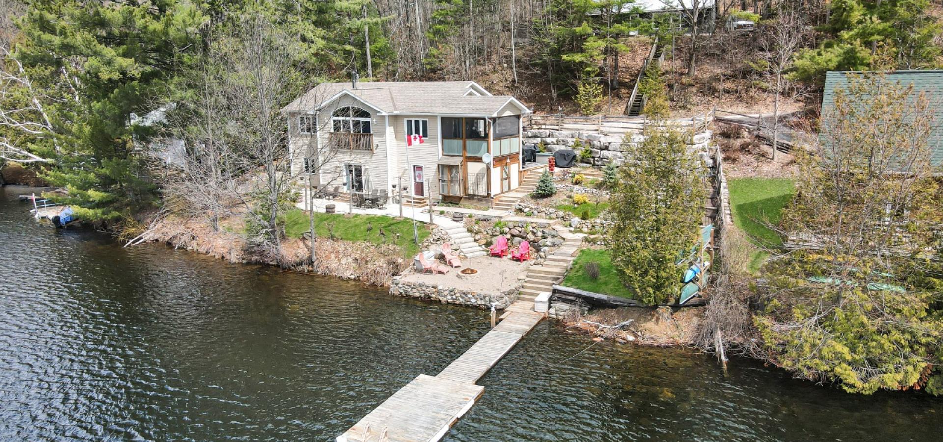 white cottage with landscaping to the lake, dock in the lake, and a cottage up behind