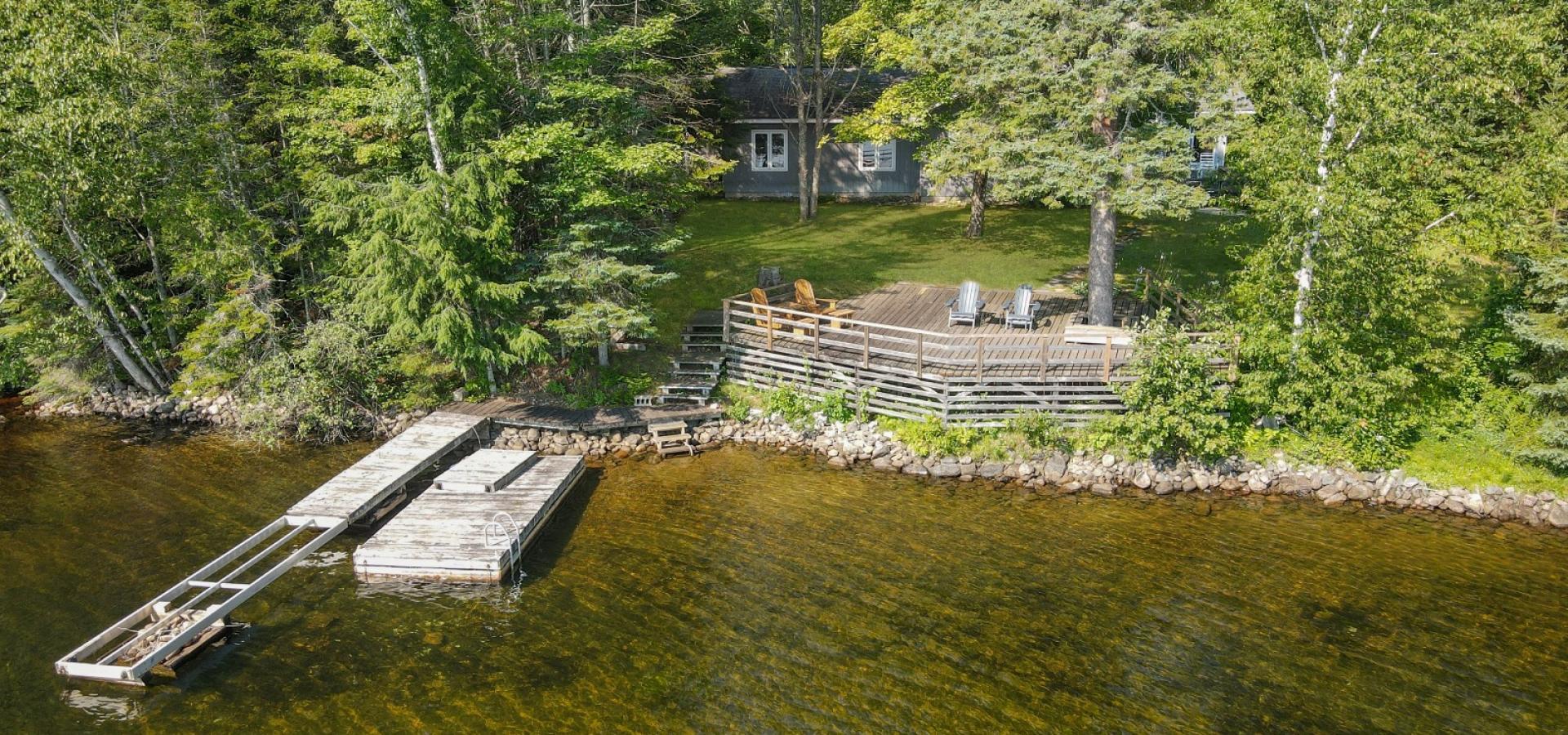 grey cottage hidden behind trees, green grass, large lake side deck and dock in the lake.