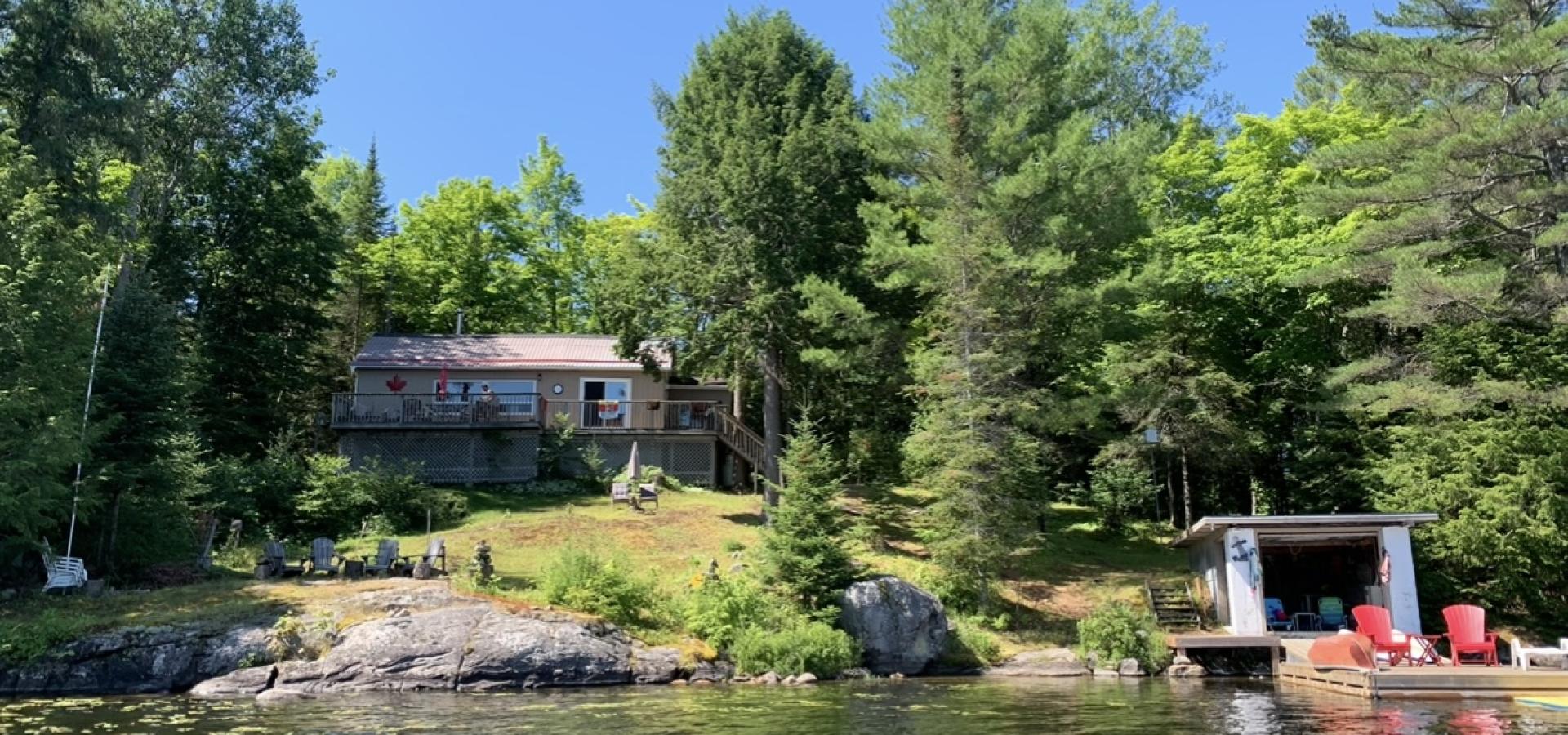 Cottage from the lake with trees, a boat house and 2 red chairs