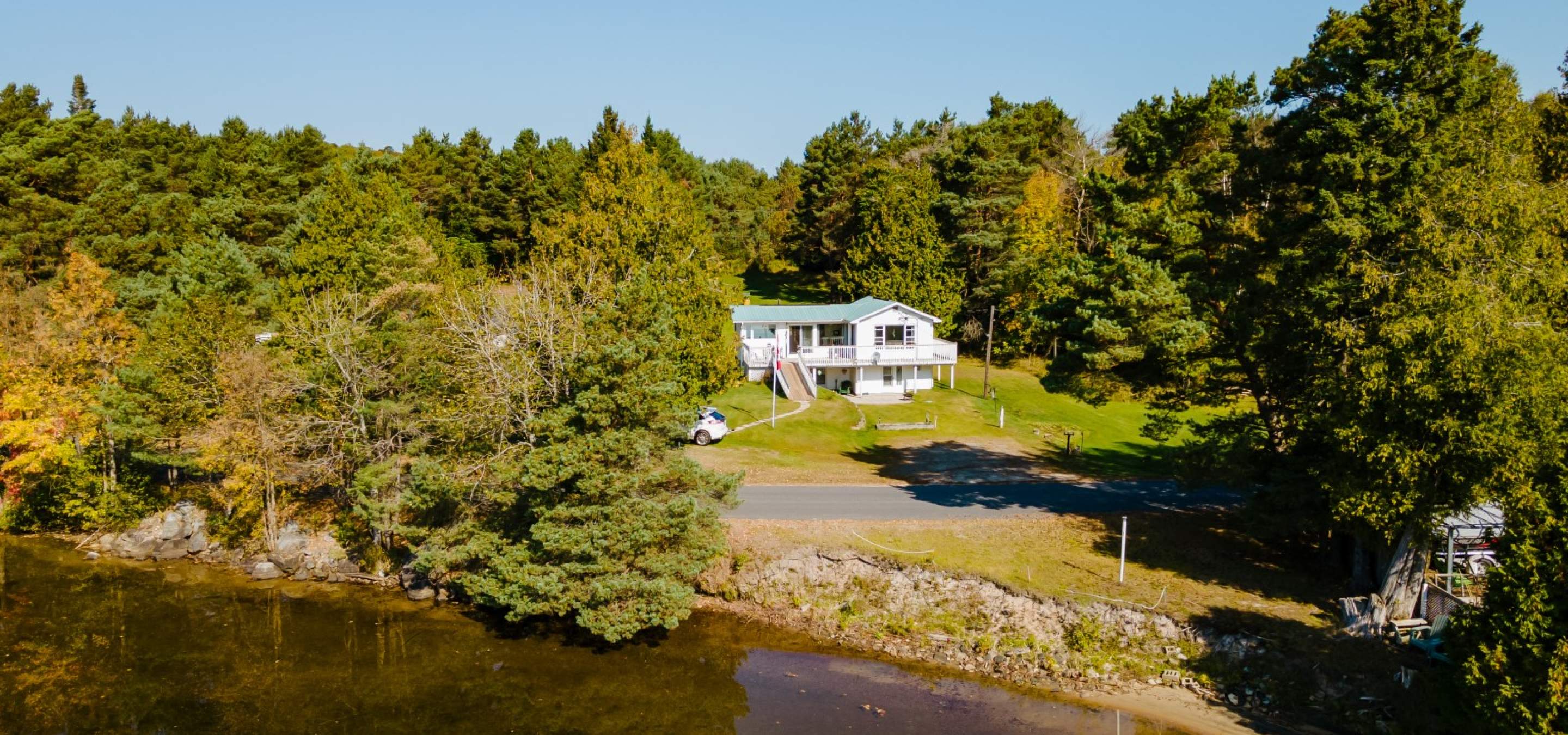 White house with trees surrounding it, grass out front with a road between cottage and lake. Beach front. 