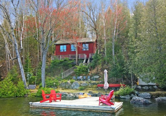 Red cottage with 3 windows, surrounded by trees. Dock in front with 4 red chairs