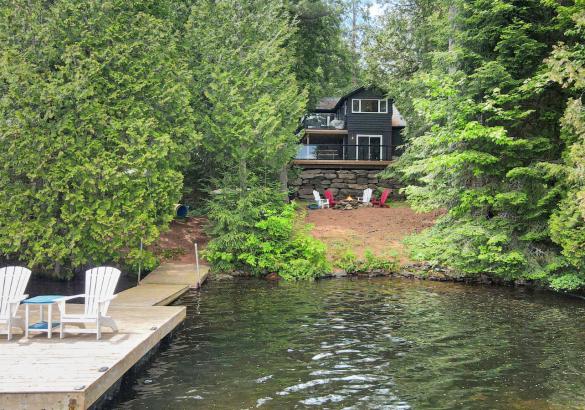 Cottage behind trees with a dock and 2 white muskoka chairs