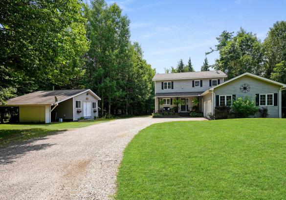 Grey house with attached garage, green grass, gravel laneway, and detached shop