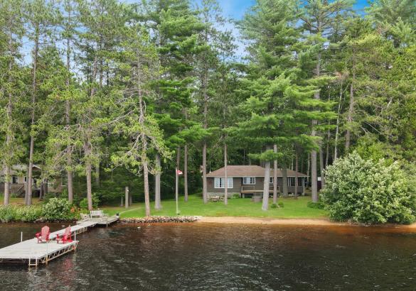 Grey cottage behind trees, sand beach and long dock with 2 red chairs