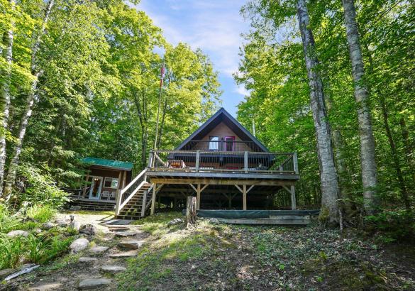 Chalet style cottage with large front deck, bunkie to the left and surrounded by trees