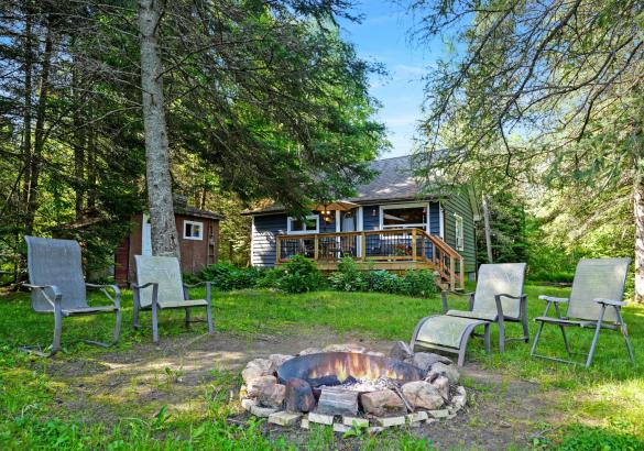 cabin with trees, a fire pit in front with 4 chairs around it.