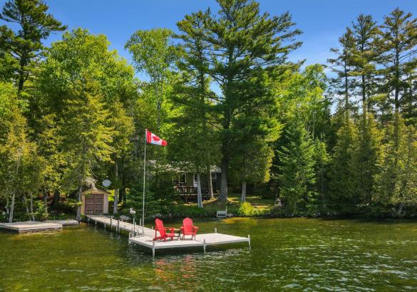 Cottage hidden behind trees, 2 red muskoka chairs on a dock with canada flag.