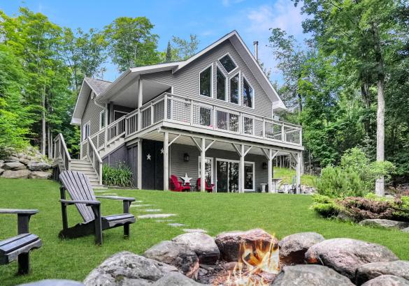 grey chalet style house with wrap around upper deck, and firepith with 2 muskoka chairs