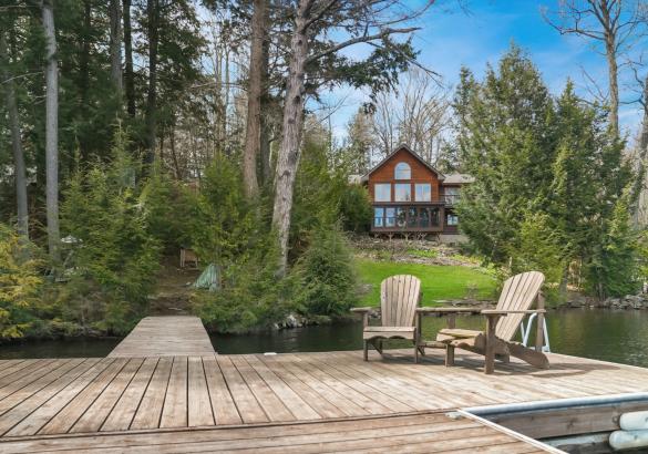 Cottage in the background with a lot of windows, surrounded by trees and a dock in front with 2 Muskoka chairs.