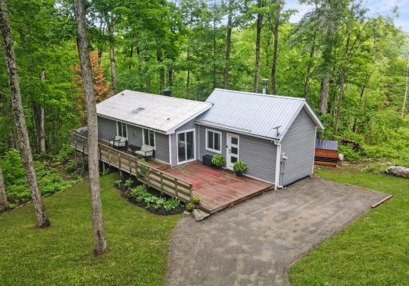 Grey home with white roof, doors and windows. Large front deck, gravel laneway and grass.