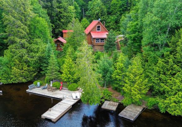 View of a cottage from the lake, surrounded by trees, with a cottage that has a red roof