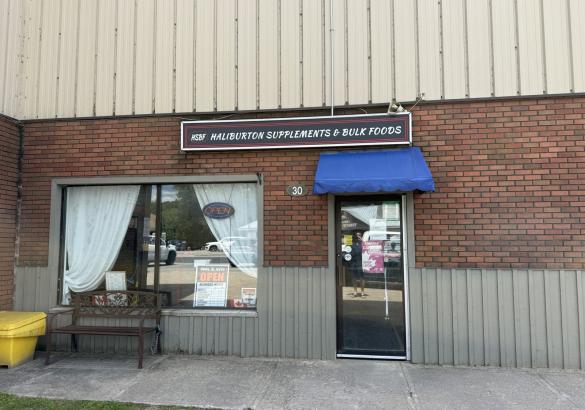 Brick store front with large window, glass door with blue awning. Business sign reading Haliburton Supplements and Bulk Foods